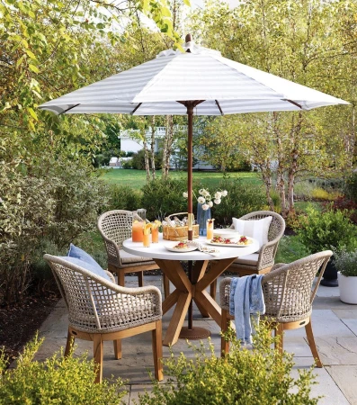 Outdoor dining setting with a round marble table, wicker chairs, striped umbrella, and drinks on a stone patio surrounded by green foliage.