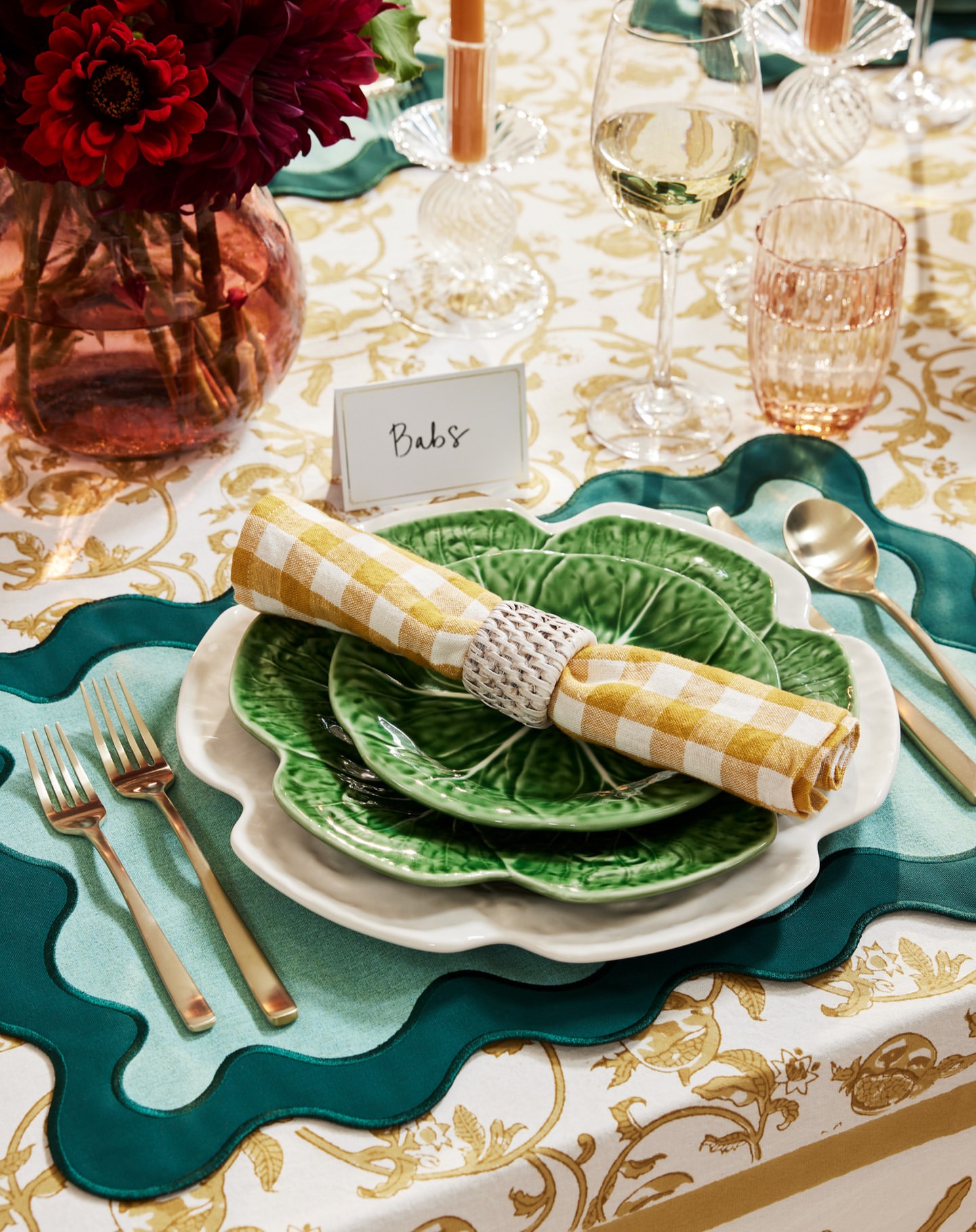 Table setting with flowers, candles, green and white plates, a yellow gingham napkin, silverware, and glassware on a patterned tablecloth.