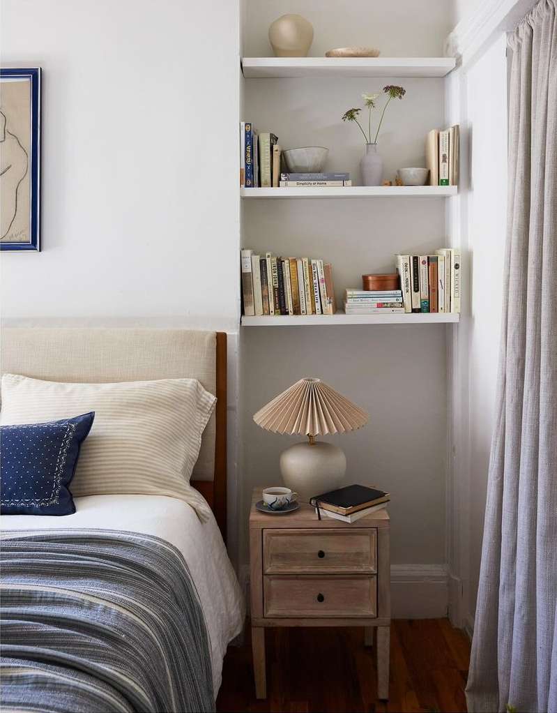 Cozy bedroom interior with a neatly made bed, a bedside table with a lamp, and shelves filled with books and decorative objects.