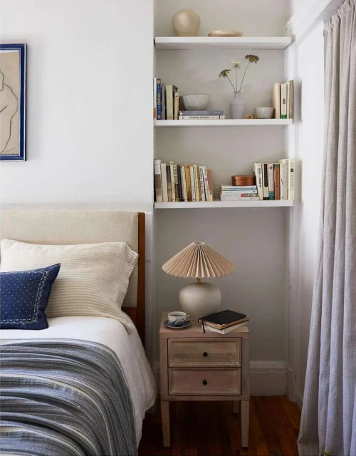 Cozy bedroom interior with a neatly made bed, a bedside table with a lamp, and shelves filled with books and decorative objects.