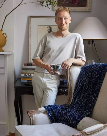 Adult male with red hair holding a striped teacup in an interior living room with furniture, books, and artwork on the wall.