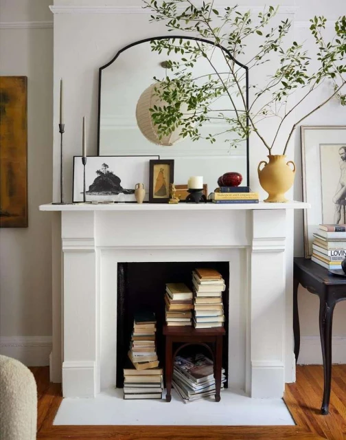 Interior of a living room with a white fireplace mantel decorated with books, artwork, candles, a mirror, and green leafy branches.
