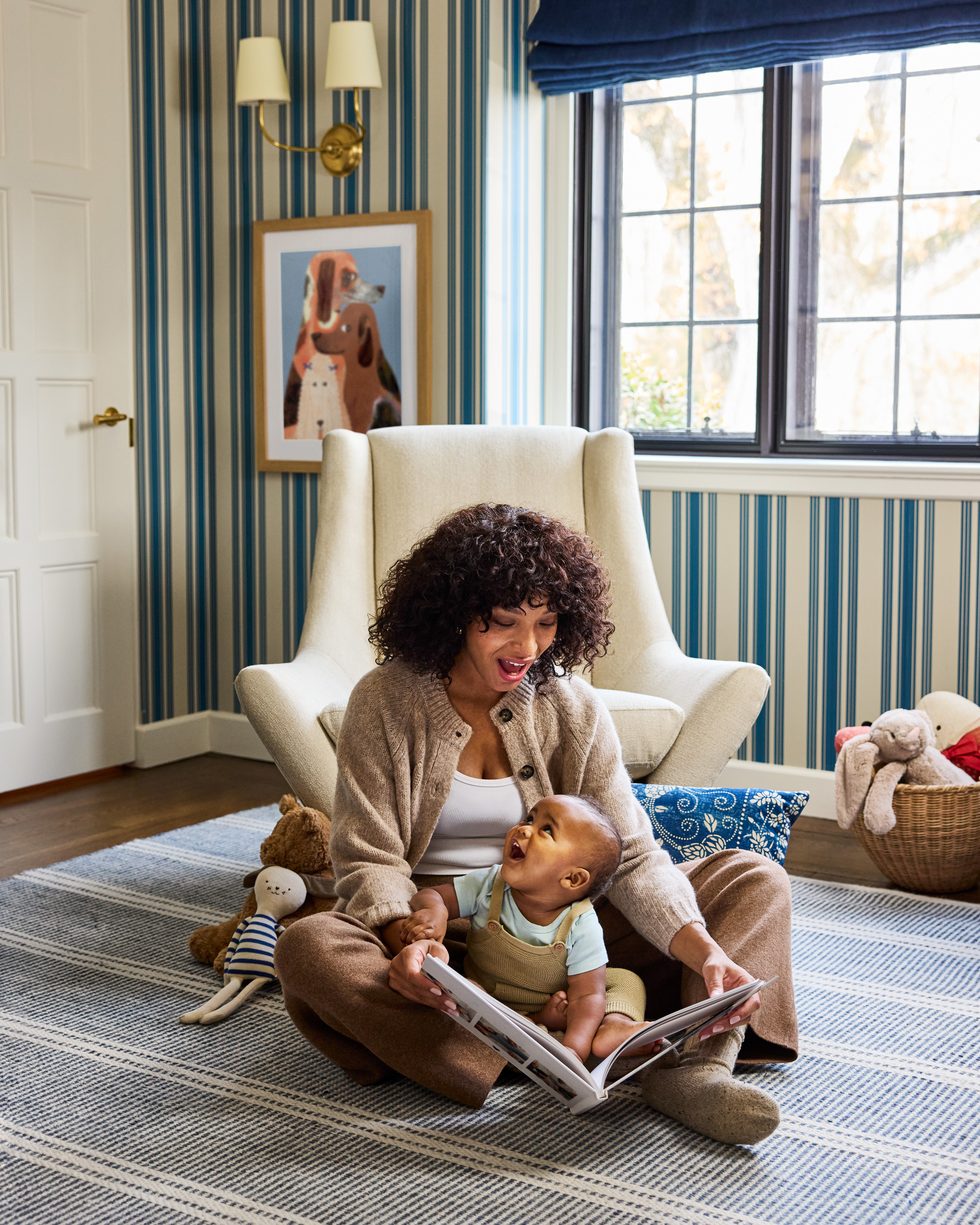 A smiling Black mother with curly hair reads a book to her baby sitting on the floor of a nursery with striped wallpaper.