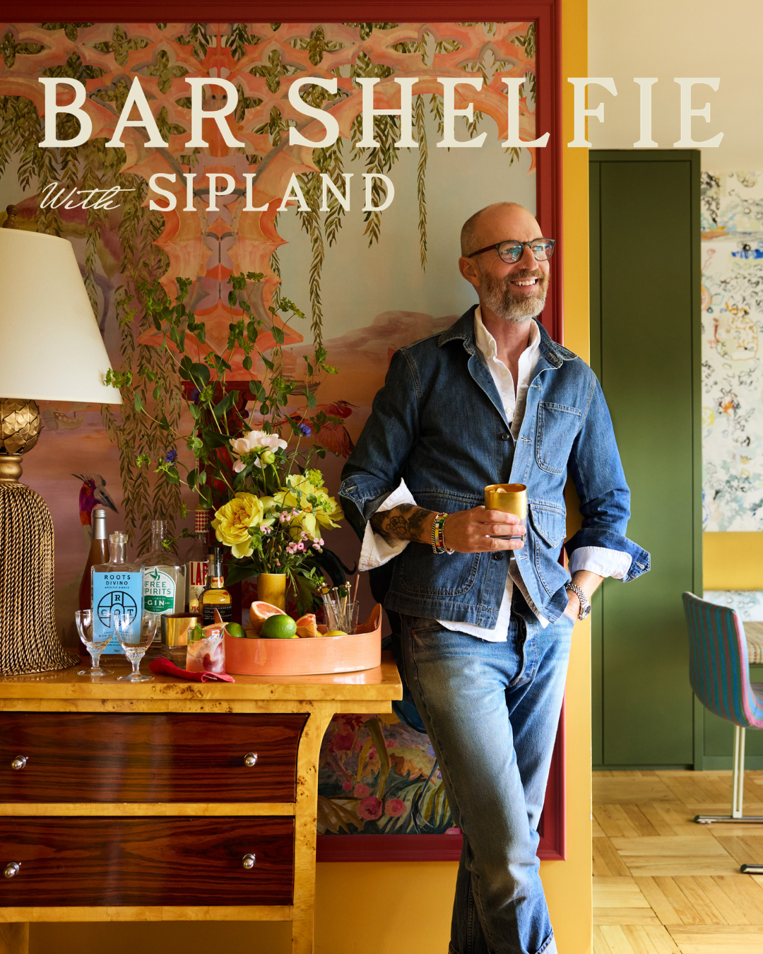 Man with glasses and denim jacket holding a gold tumbler beside a colorful home bar display in a modern room.