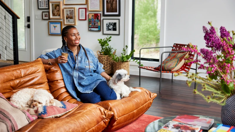 A Black woman with braided hair sits on a cognac leather sofa holding a small cup, with two small dogs resting beside her in a bright living room.