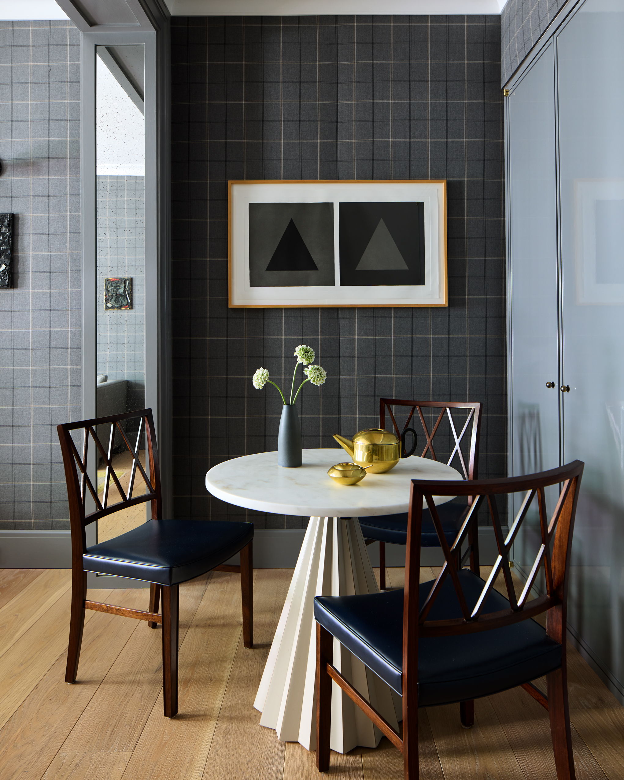 A round white marble table sits on a fluted pedestal base in a room with dark grey plaid wallpaper and blue upholstered wooden chairs.