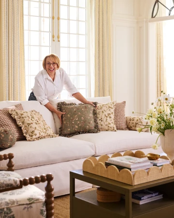 Smiling woman with glasses in a white shirt arranges decorative floral-patterned ruffled pillows on a cream-colored couch in a bright room.