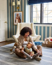 Woman and baby sitting on a rug reading a book in a living room with striped wallpaper and an armchair