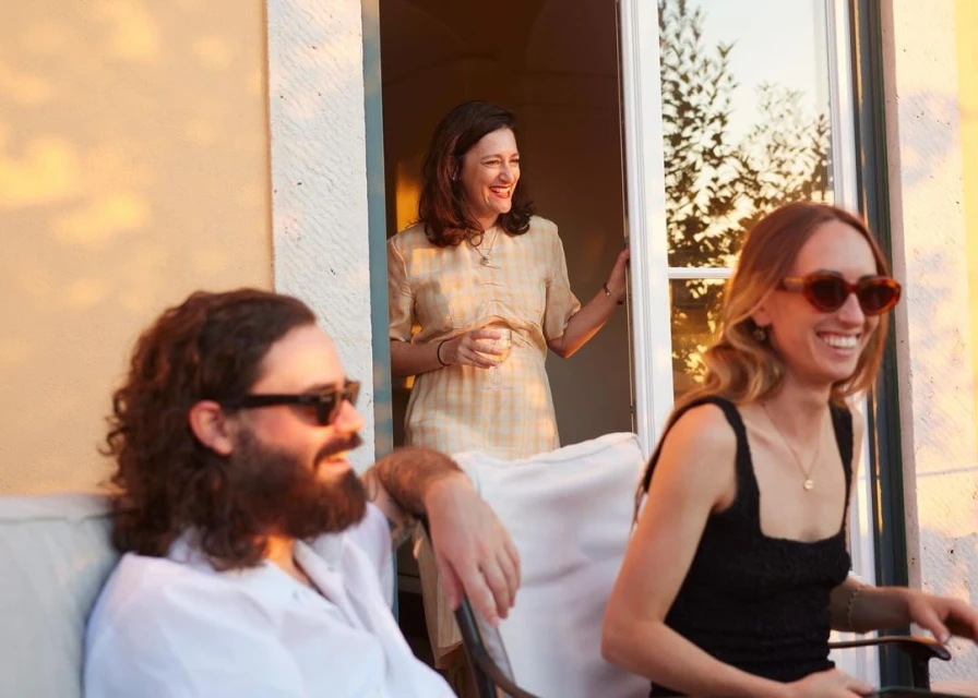 Three adults are outside a building with white trim, enjoying time on a patio; two are seated, and one woman is standing in the doorway.