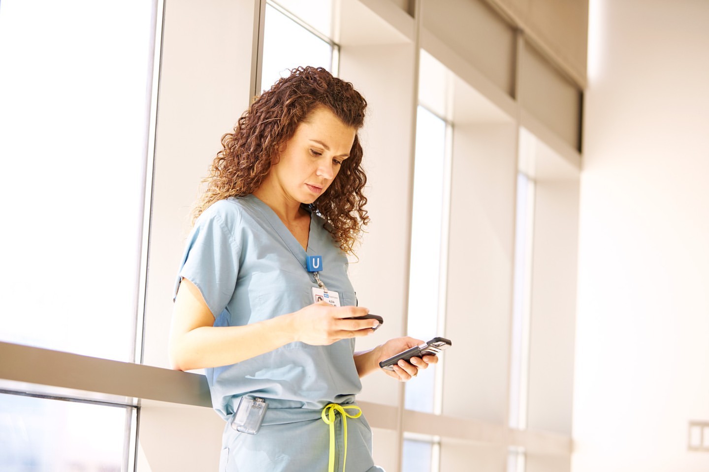 Adult female healthcare worker in scrubs checks two smartphones near large window with natural light in a hospital corridor.