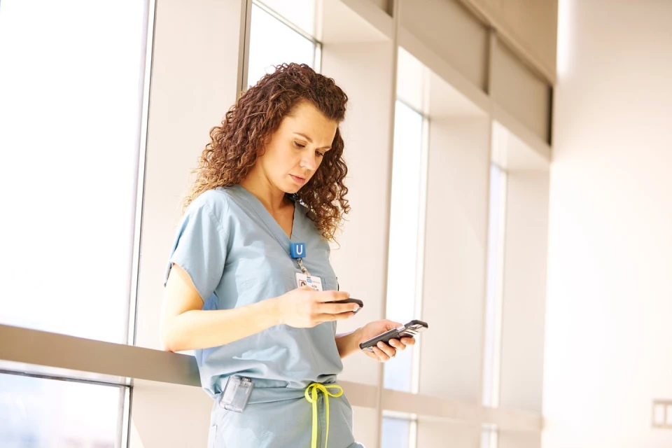 Adult female healthcare worker in scrubs checks two smartphones near large window with natural light in a hospital corridor.