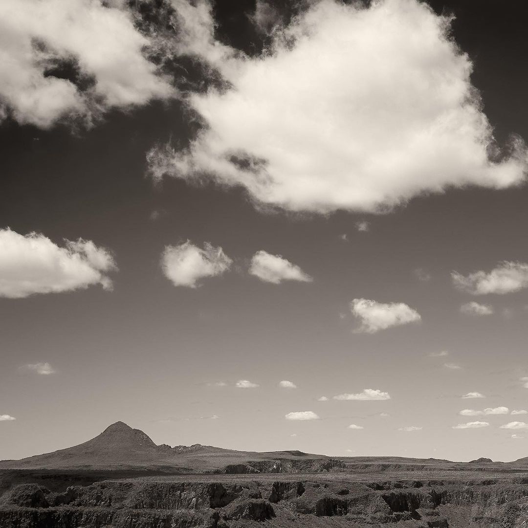 Monochrome landscape featuring a small mountain range under a cloudy sky. The monochromatic tones emphasize the texture of the clouds.