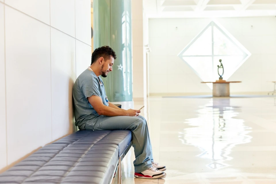 Healthcare professional in scrubs seated on bench using a smartphone inside a bright, modern hospital hallway with natural light.