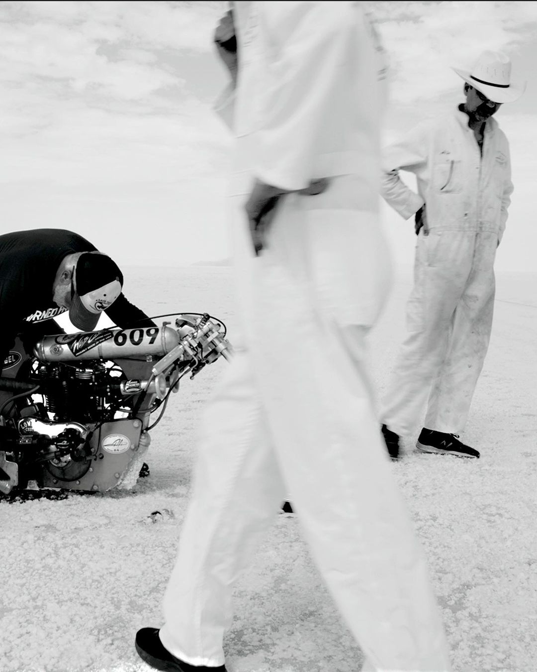 Black and white shot of two people near a race car, one person is working on the car in a desolate landscape under an overcast sky.