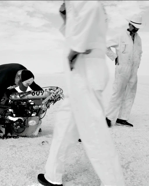 Black and white shot of two people near a race car, one person is working on the car in a desolate landscape under an overcast sky.