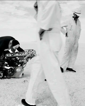 Black and white shot of two people near a race car, one person is working on the car in a desolate landscape under an overcast sky.