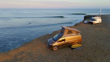 Camper van parked on a cliff overlooking the ocean at sunset, with a surfboard nearby, other camper vans in the background.