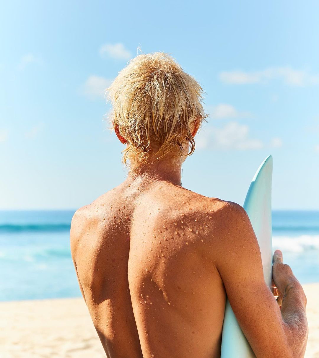 Blonde surfer holding a surfboard on a sandy beach looking out at the ocean under a clear blue sky