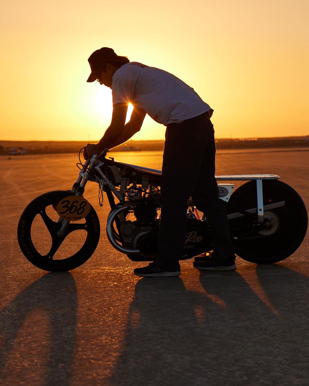 A man with long hair is leaning over a vintage motorcycle at sunset on a flat surface, with a sunlit sky in the background.