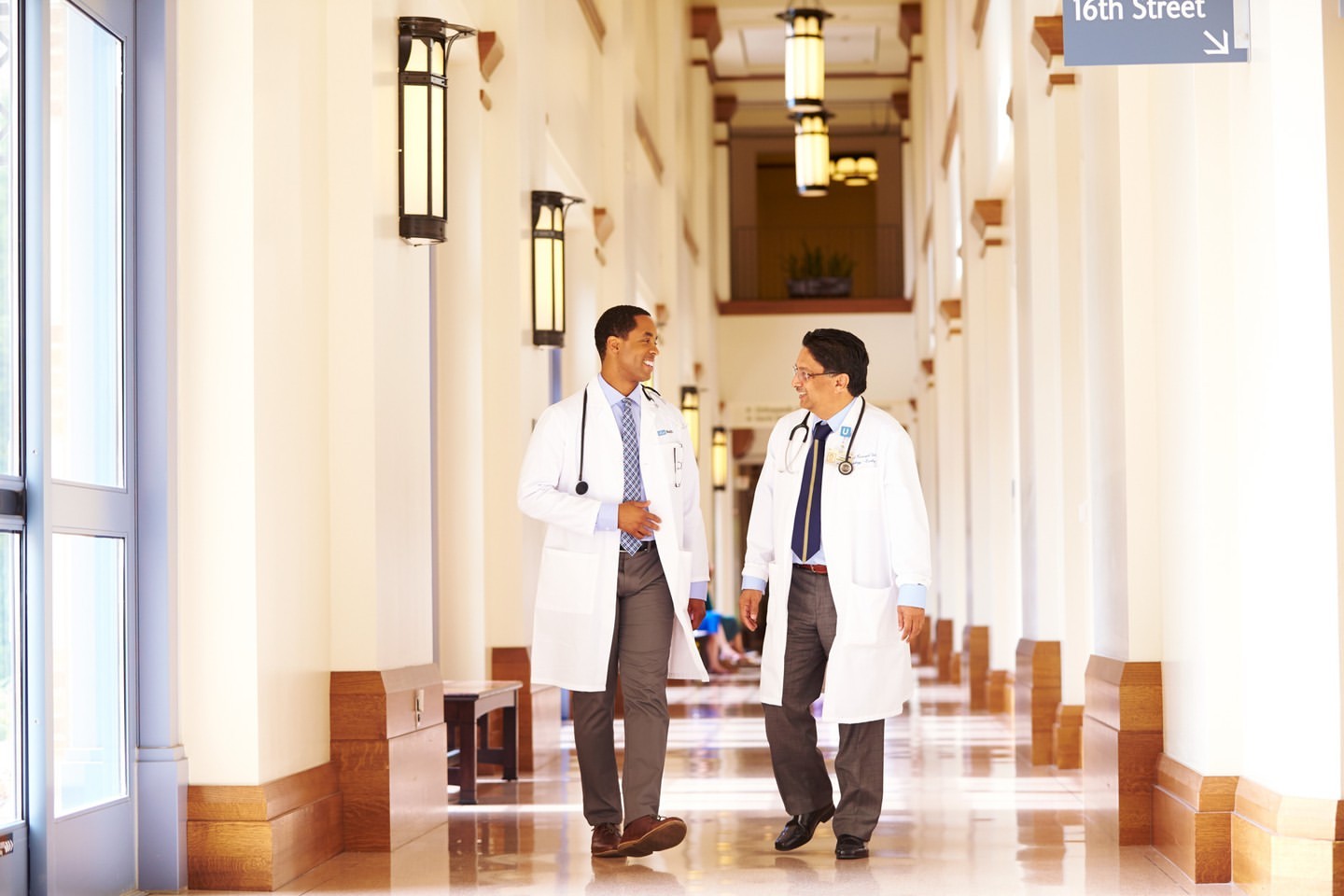 Two doctors in white coats walk and converse in a well-lit hallway of a medical facility, with a sign pointing to 16th Street.