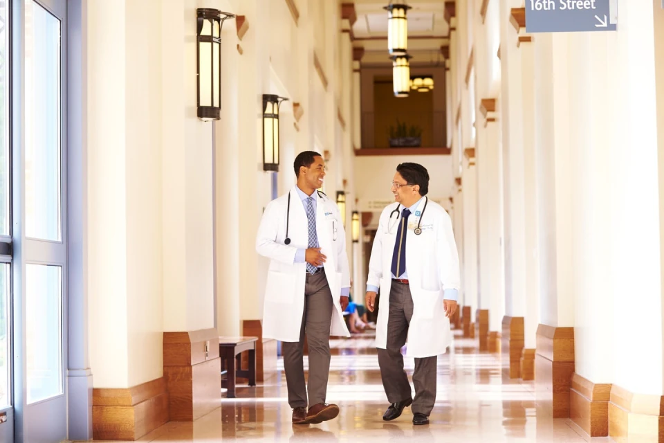Two doctors in white coats walk and converse in a well-lit hallway of a medical facility, with a sign pointing to 16th Street.