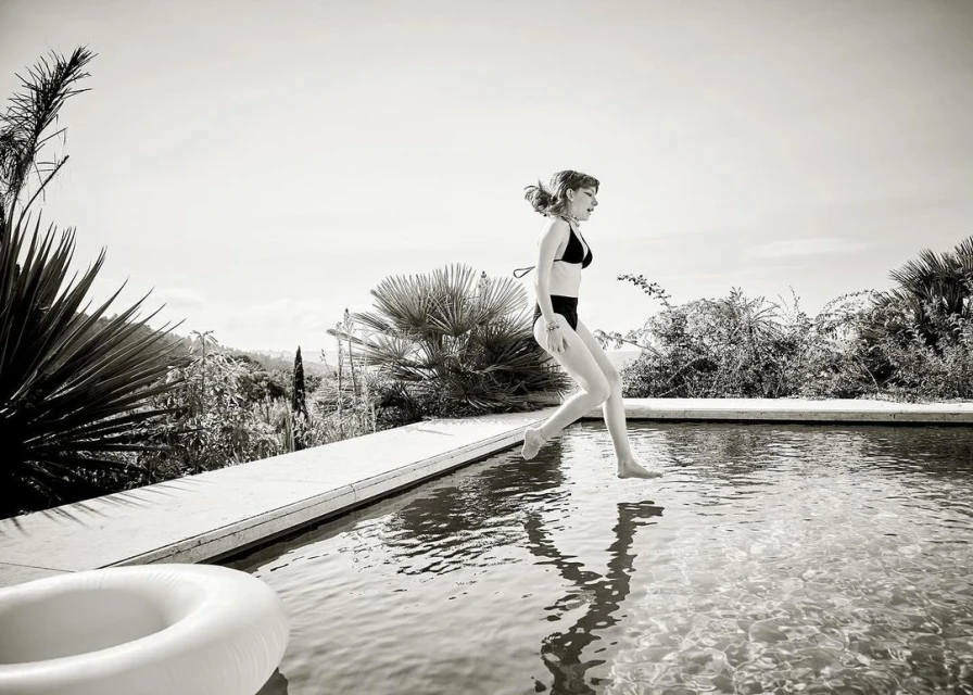 A young woman wearing a black bikini jumps into an outdoor pool, surrounded by greenery, captured in black and white with natural lighting.