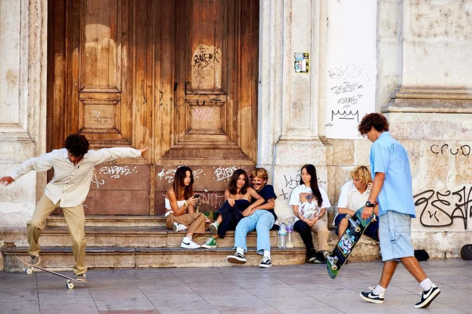 Group of young adults socializing outdoors; one man on a skateboard, and others sitting on the steps of a building with graffiti.