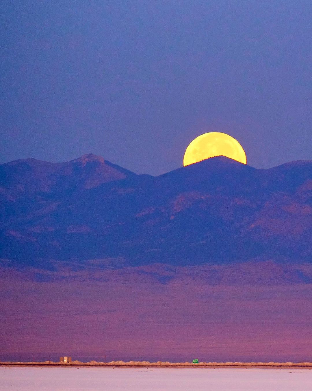 Full moon rising over mountain range at dusk creating a scenic landscape view with blue sky in background and flat, textured land in foreground.
