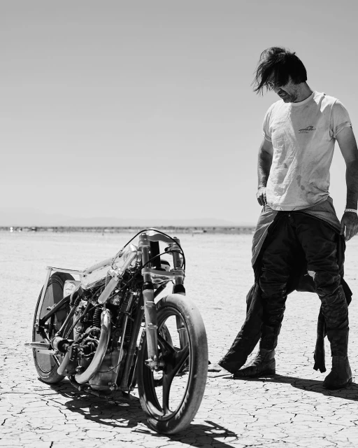 Monochrome shot of a man in a t-shirt standing next to a custom-built motorcycle on a cracked desert surface under a clear sky.