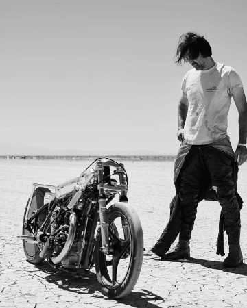 Monochrome shot of a man in a t-shirt standing next to a custom-built motorcycle on a cracked desert surface under a clear sky.