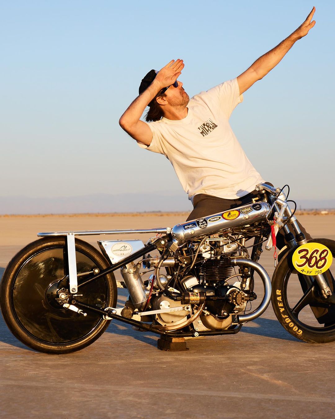 Adult male poses with a custom race motorcycle on a flat desert landscape with a clear sky during daytime, showcasing design and engineering.