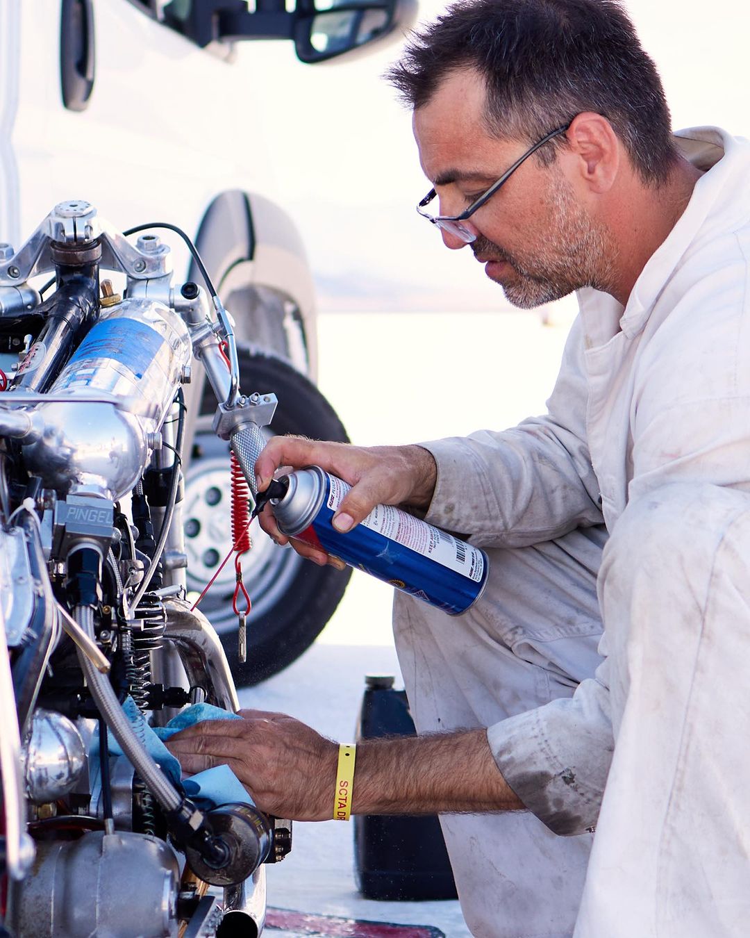 Adult male sprays lubricant on the engine of a vintage motorcycle outdoors during daytime, wearing glasses and a white jumpsuit.