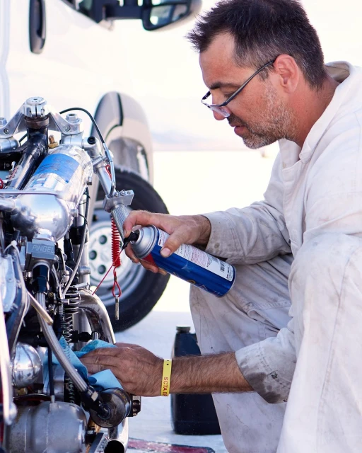 Adult male sprays lubricant on the engine of a vintage motorcycle outdoors during daytime, wearing glasses and a white jumpsuit.