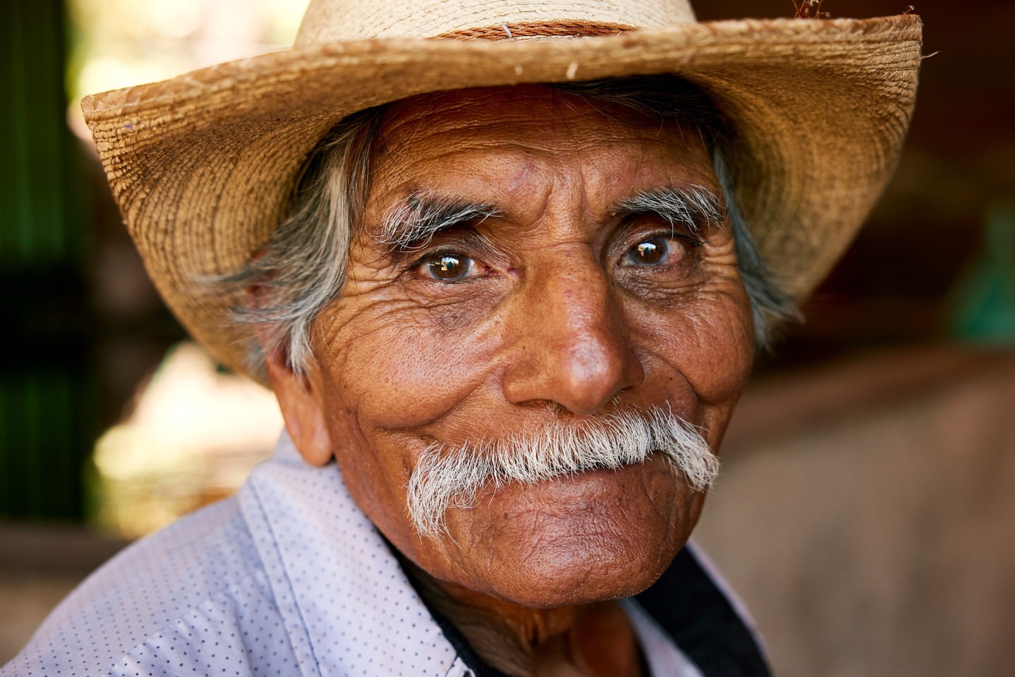 Close-up portrait of a smiling, elderly Hispanic man with a mustache, wearing a straw hat, looking directly at the camera.