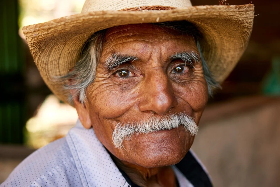 Close-up portrait of a smiling, elderly Hispanic man with a mustache, wearing a straw hat, looking directly at the camera.