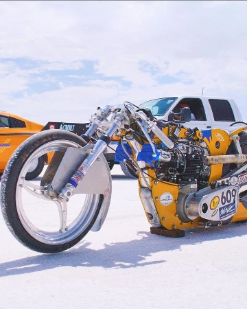 Custom-built motorcycle with yellow and blue accents stands out against a white salt flat, flanked by support vehicles, under a cloudy sky.