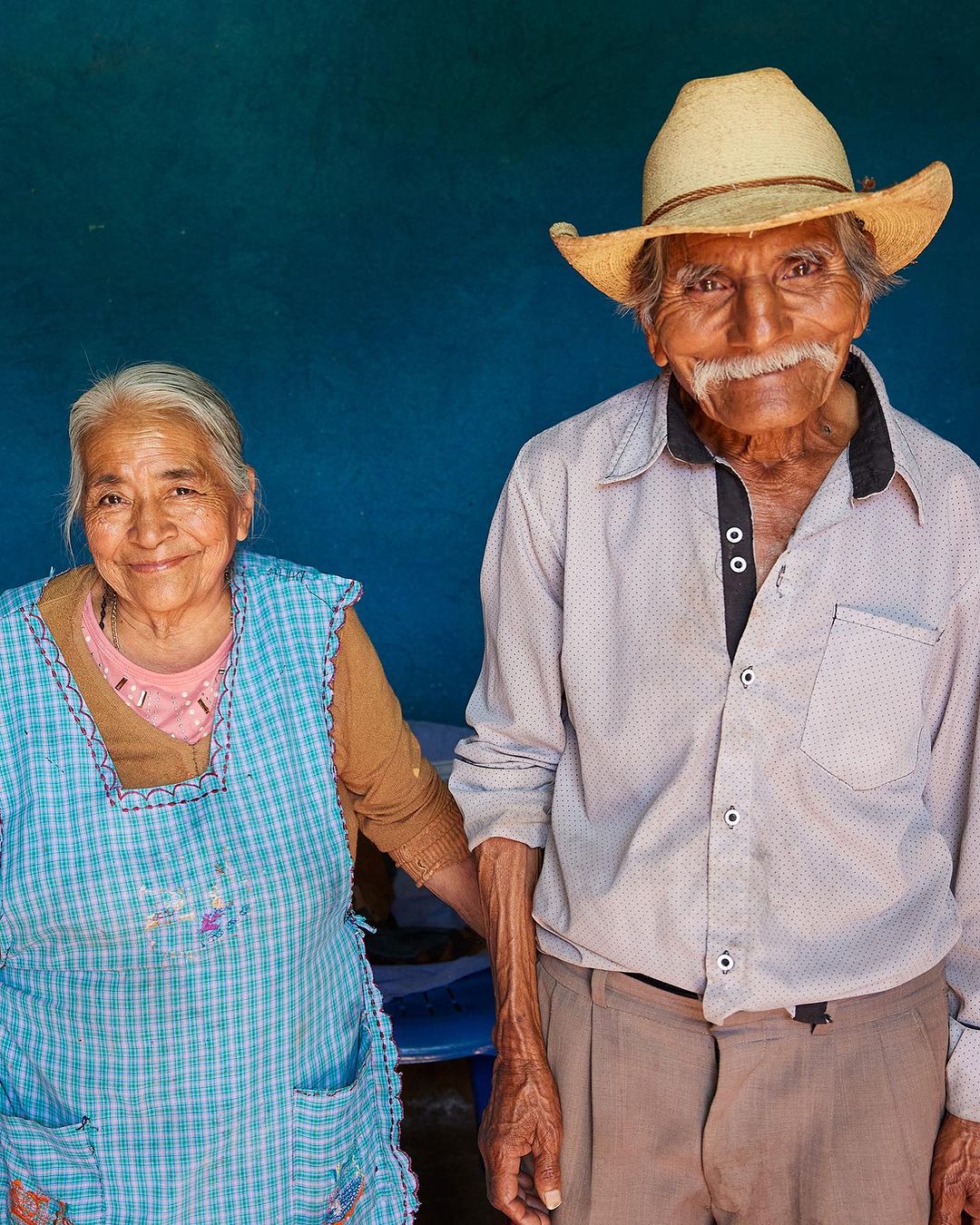 Elderly couple posing indoors against a blue wall, the man is wearing a straw hat and the woman is wearing a blue apron.