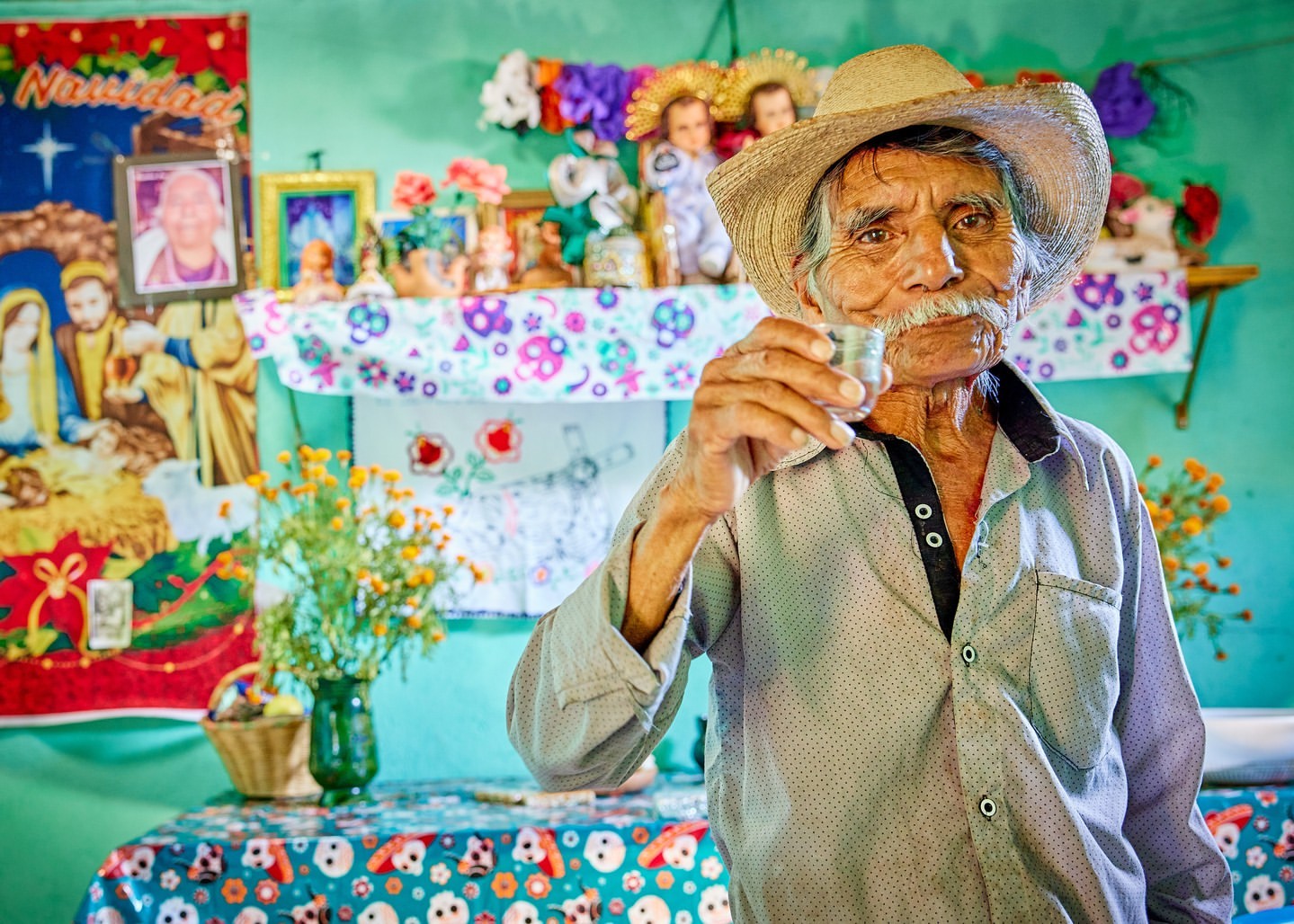 Elderly man in straw hat holds drink in a glass, posing indoors with cultural and religious decorations on a blue-green wall.