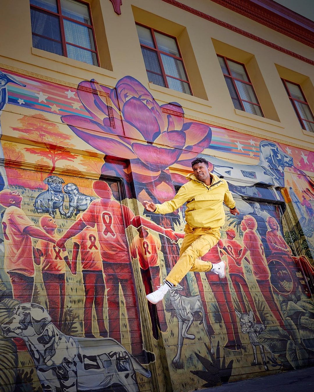 Man in yellow tracksuit jumps in front of large outdoor mural featuring stylized figures, animals, and colorful artwork on a building.
