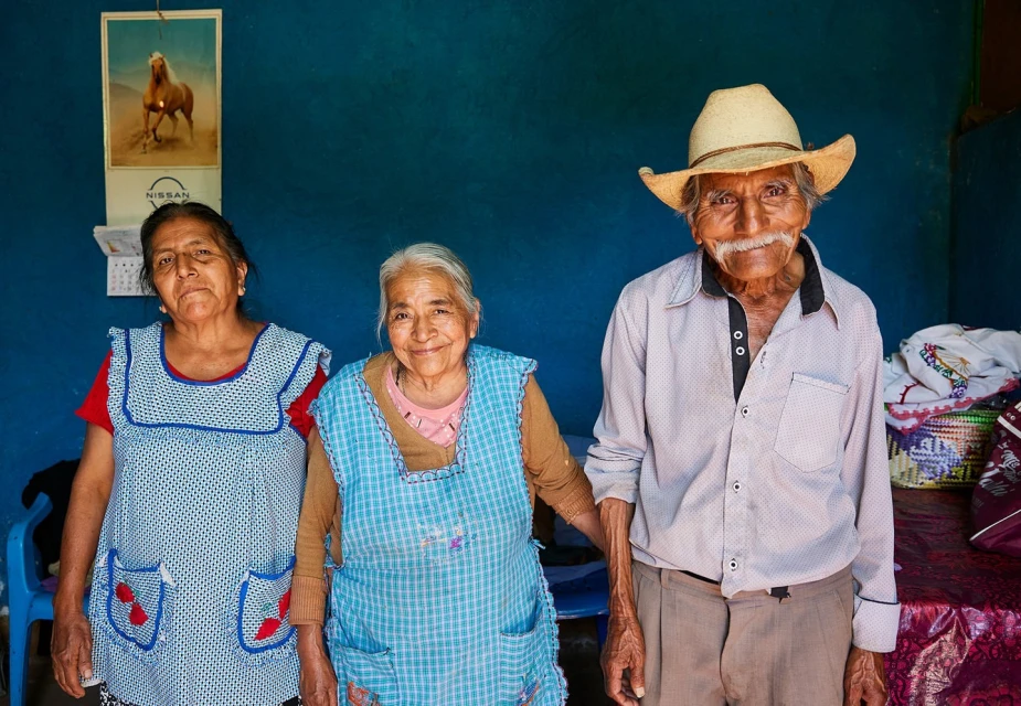 Portrait of three elderly people standing indoors: an elderly man in a straw hat, and two elderly women in aprons with a blue wall backdrop.
