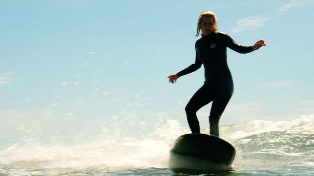 A woman in a wetsuit is surfing on an object in the ocean with a blue sky. She is standing with her arms out for balance.