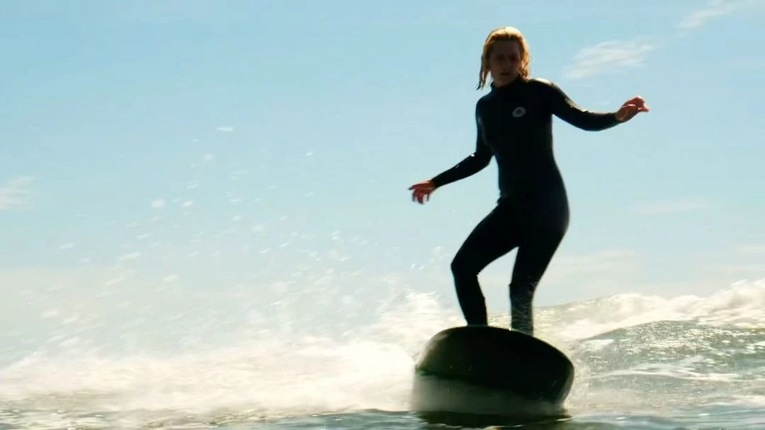 A woman in a wetsuit is surfing on an object in the ocean with a blue sky. She is standing with her arms out for balance.