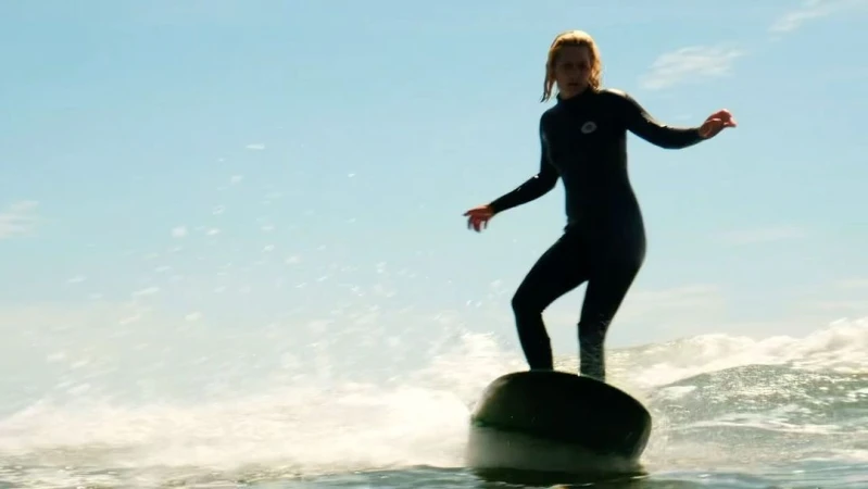 A woman in a wetsuit is surfing on an object in the ocean with a blue sky. She is standing with her arms out for balance.