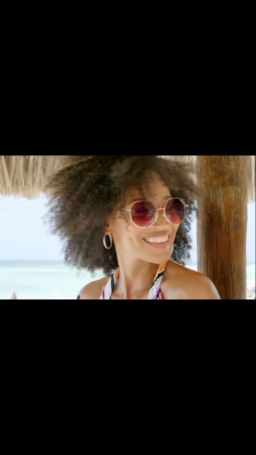 Smiling woman with afro and round sunglasses relaxing under a thatched roof structure, with the beach and ocean visible in the background.