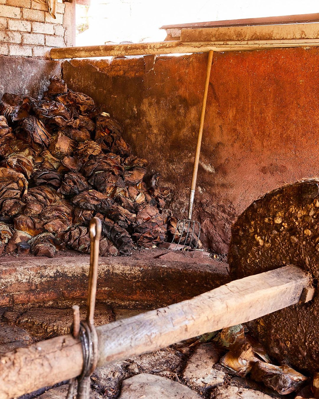 Agave plants piled next to a stone grinding wheel indoors, ready for processing into tequila or mezcal in a rustic distillery.