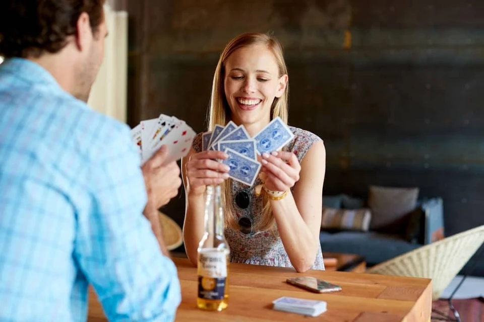 Woman holding playing cards while playing with a man at a wooden table with a bottle of beer and a cell phone in an indoor setting.