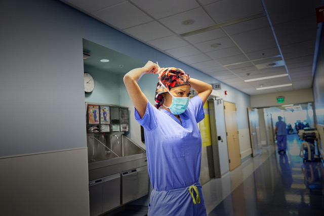 Healthcare worker in blue scrubs and mask adjusts head covering in a brightly lit hospital hallway near a stainless-steel sink.