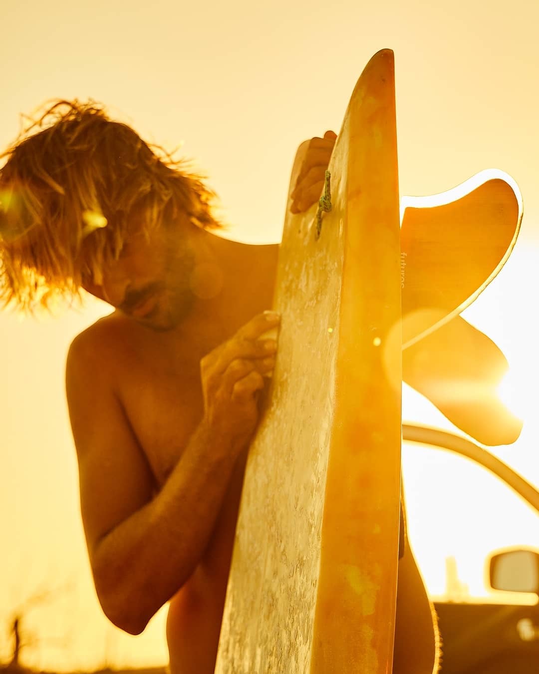 Male surfer standing with a surfboard at sunset. The sunlight creates a warm, golden glow around the man and his board at dusk.