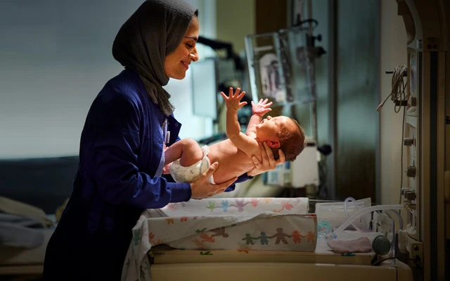 A caring healthcare worker in a hijab holds a newborn baby in a hospital room, both smiling, with medical equipment in background.