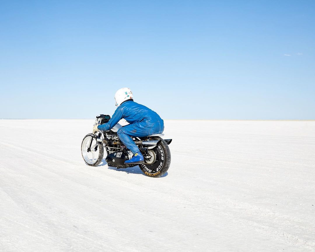 Motorcycle racer in blue suit speeds across a white salt flat under a clear blue sky. Action shot from the rear.